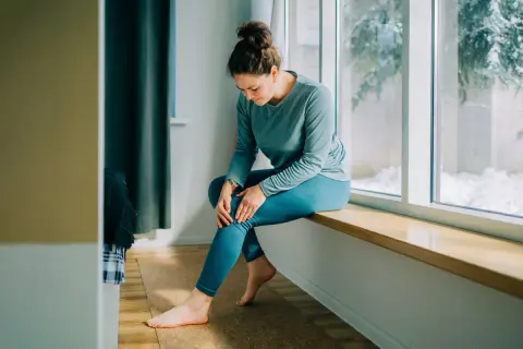 Femme assise près d'une fenêtre, tenant son genou, possiblement à cause d'une **durée courbature** après un effort.