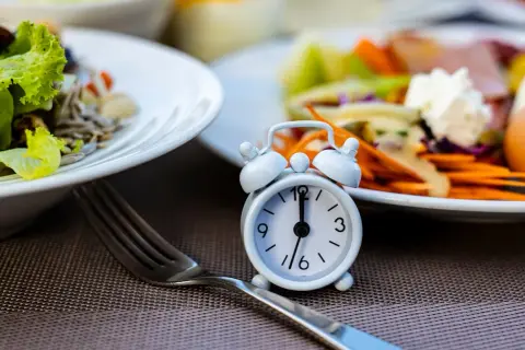 Réveil miniature sur une table, symbolisant le jeûne intermittent 12h. Des assiettes de salade et de légumes sont floues en arrière-plan.