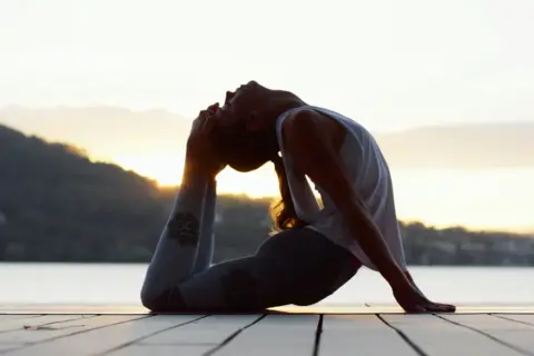 Silhouette d'une femme en posture de yoga au bord de l'eau, le soleil se levant derrière elle. Ses leggings sont ornés de symboles, évoquant les mantra.