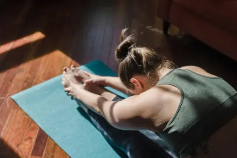 Une femme fait du yoga chez soi, étirant ses jambes sur un tapis bleu, baignée par la lumière du soleil.