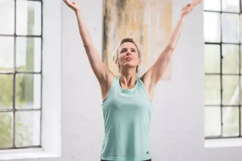 Femme aux bras levés, profitant d'un moment pour calmer son mental. La lumière naturelle baigne la pièce, accentuant sa sérénité.