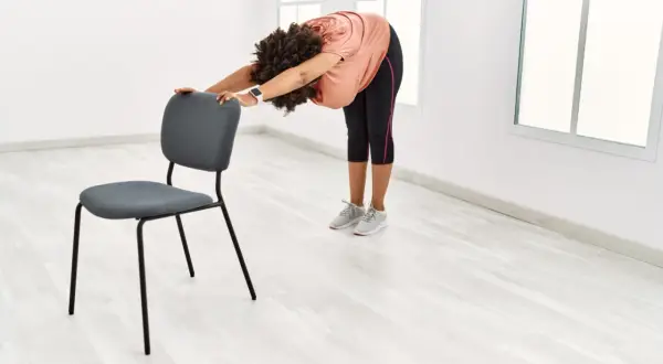 Femme pratiquant le yoga sur chaise, se penchant en avant pour étirer son dos, mains posées sur le dossier du siège.
