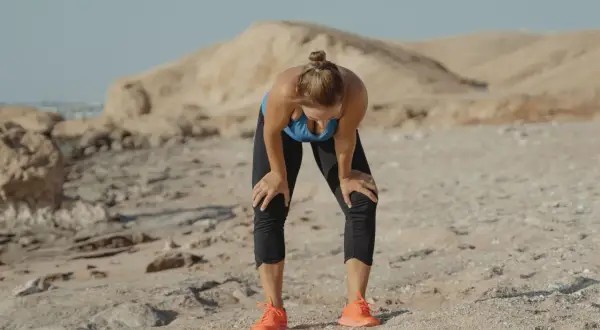 Une femme s'arrête, penchée en avant, les mains sur les genoux, visiblement fatiguée par son sport sous la canicule. Le paysage désertique accentue la chaleur.