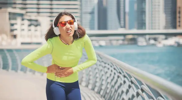 Une coureuse en tenue de sport, écouteurs aux oreilles, souffre d'un point de côté aigu en pleine course le long d'une promenade urbaine.