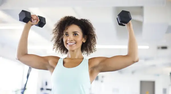 Femme souriante faisant des exercices avec haltères pour maigrir des bras. Elle est dans une salle de sport.