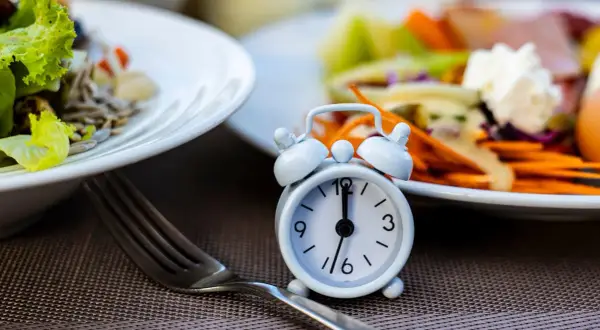 Réveil miniature sur une table, symbolisant le jeûne intermittent 12h. Des assiettes de salade et de légumes sont floues en arrière-plan.