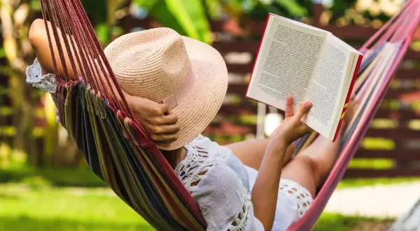 Une personne lit un livre dans un hamac coloré, profitant d'un des 7 types de repos sous un chapeau de paille.