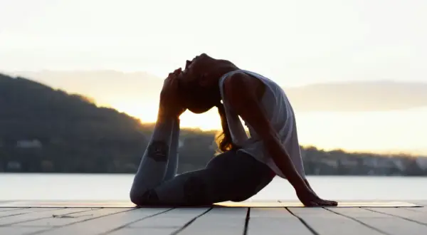 Silhouette d'une femme en posture de yoga au bord de l'eau, le soleil se levant derrière elle. Ses leggings sont ornés de symboles, évoquant les mantra.