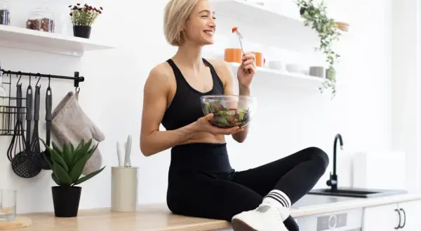 Une femme souriante en tenue de sport mange une salade fraîche. Elle savoure son repas sain dans une cuisine lumineuse.