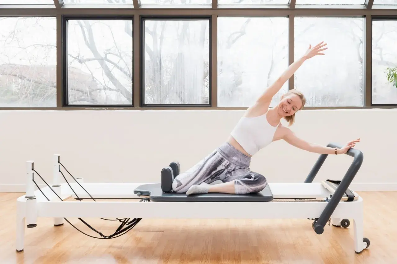 Femme souriante en plein &eacute;tat de flow sur un reformer Pilates, bras lev&eacute;s, &eacute;tirant son corps avec gr&acirc;ce.
